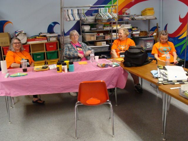 Four people sit at tables in a classroom, with pink tablecloths and craft supplies spread out.