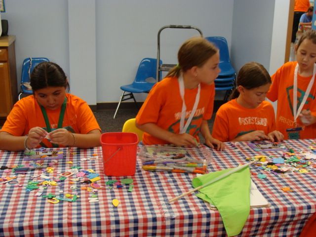 Children in orange shirts making crafts at a checkered table with colorful supplies