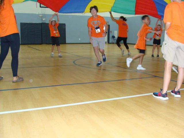 Children in orange shirts playing under a colorful parachute in a gymnasium