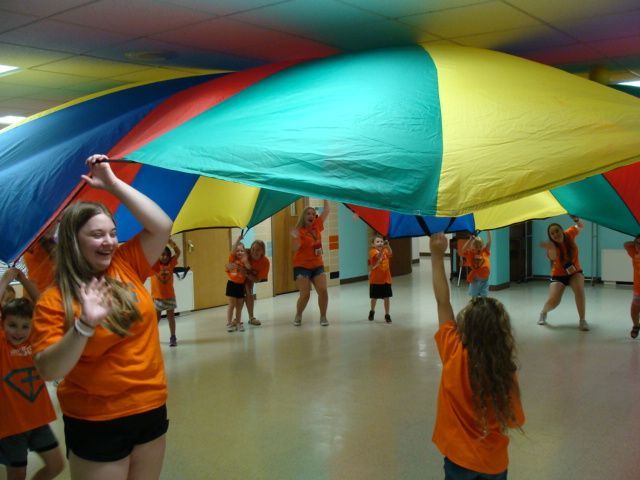 Children in orange shirts holding a colorful parachute in a gymnasium
