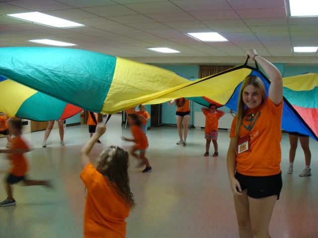 Children holding a multicolored parachute in a gymnasium, with one girl raising it overhead