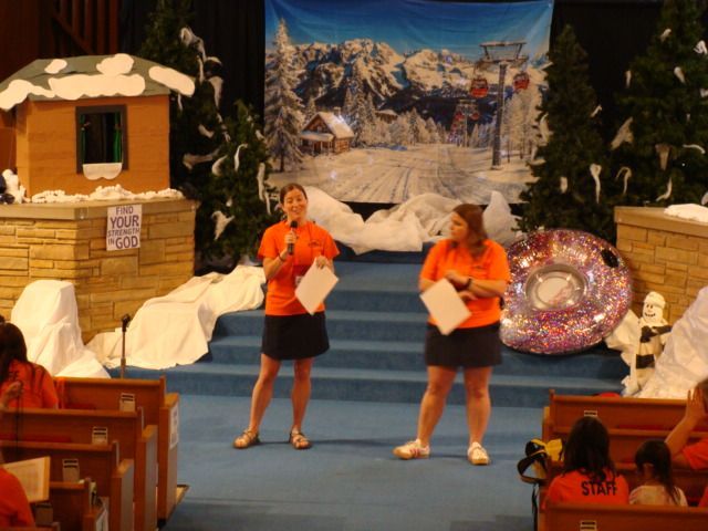 Two women on a stage with a snowy village backdrop, holding orange papers and a sparkly silver ring prop.