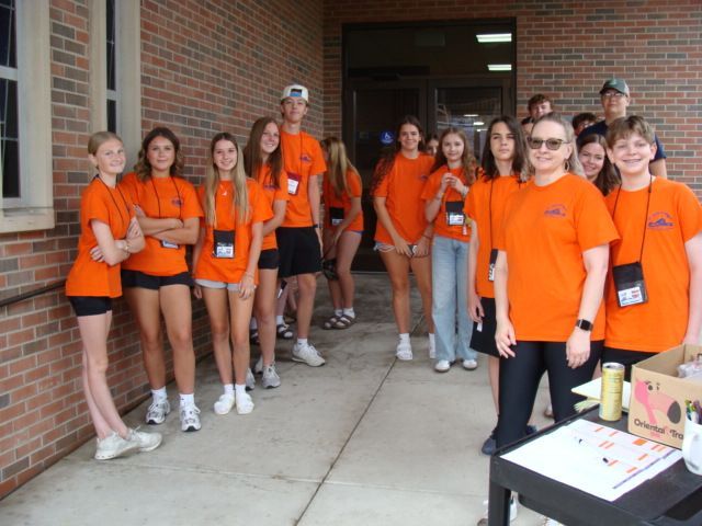 Group of people in orange shirts standing outside a brick building near a table with supplies