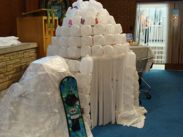 White igloo-like sculpture made of stacked plastic jugs in an indoor lobby, with a snowboard leaning against it