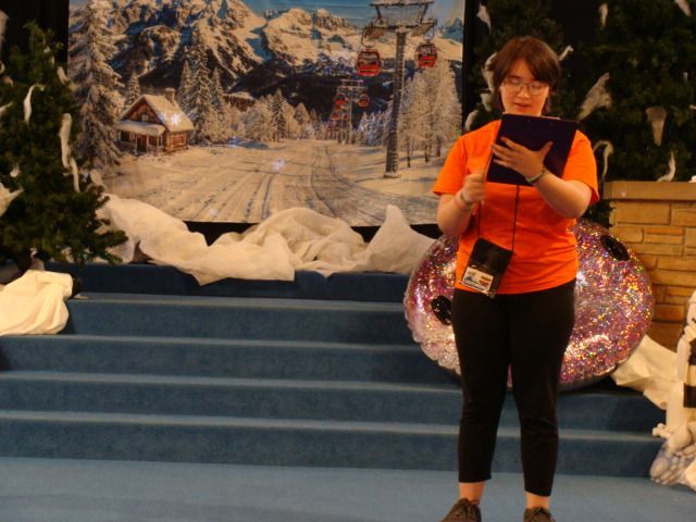 Person in orange shirt reading from a clipboard on a snowy stage with winter backdrop and decorations