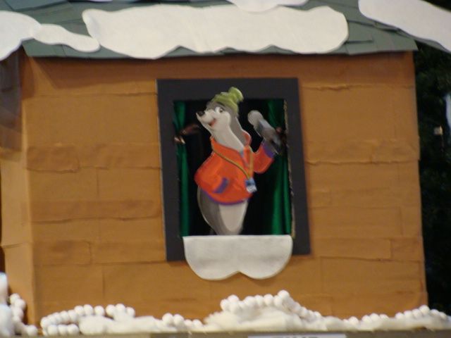 Small figurine in an orange jacket peeking from a window of a cardboard house with snow on top and at the base
