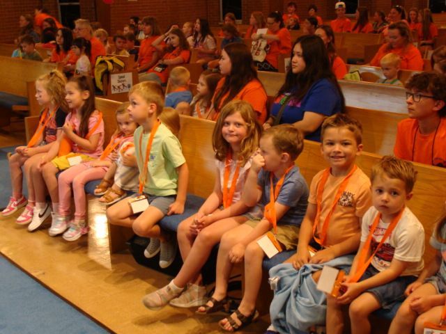 Children in orange lanyards seated in rows at an indoor event, some smiling toward the front.