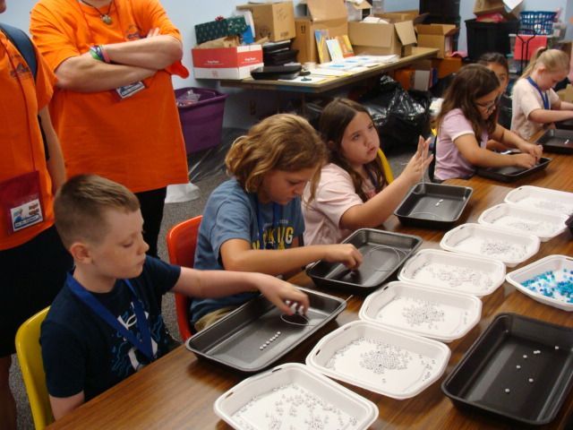 Children in a classroom serving meals into trays at a long table