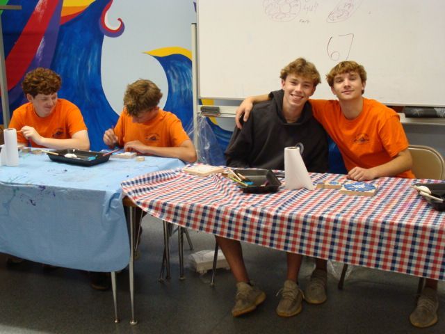 Four young people seated at two tables with red-and-white and blue tablecloths, smiling in a classroom or event room