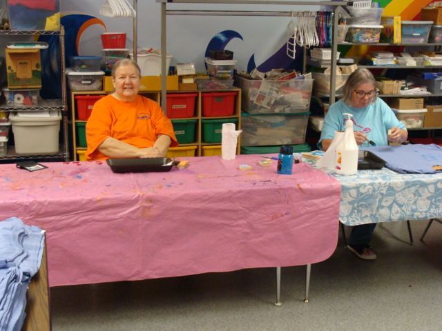 Two people seated behind tables with pink and blue tablecloths at a craft fair booth