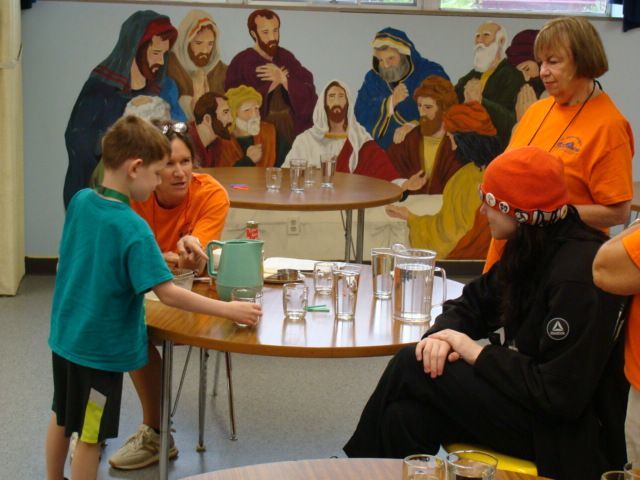 Children and adults gather around tables in a bright room, with a colorful mural in the background.