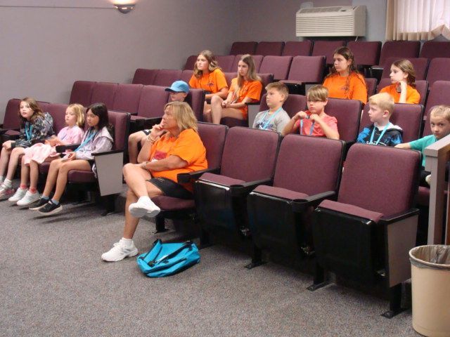 Children seated in a theater, many wearing orange shirts, watching something off-camera