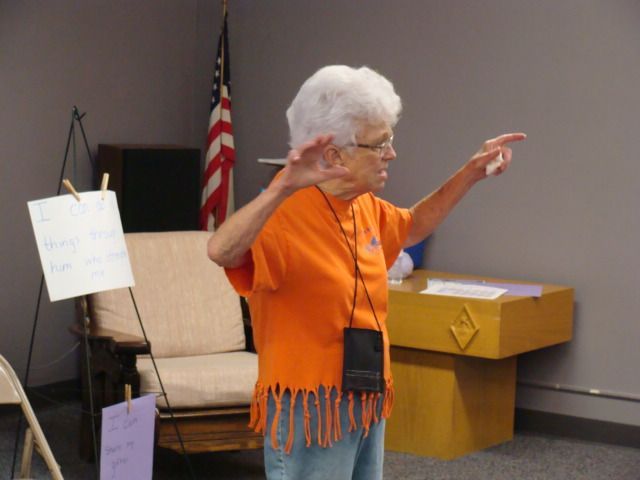 Elderly woman in orange top gesturing in a room with an American flag, chairs, and a table of boxes