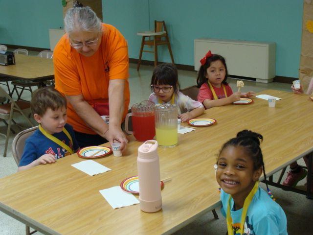 Teacher helping children with a snack table in a classroom, with plates, cups, and a smiling child in front.