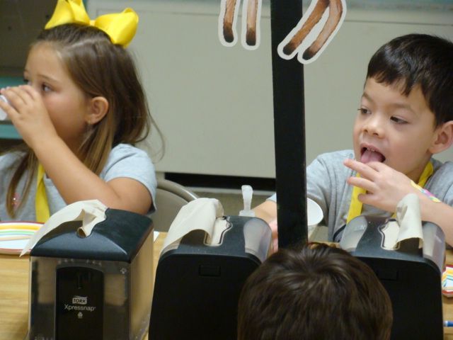 Two children seated at a table, eating snacks in a classroom-like setting.