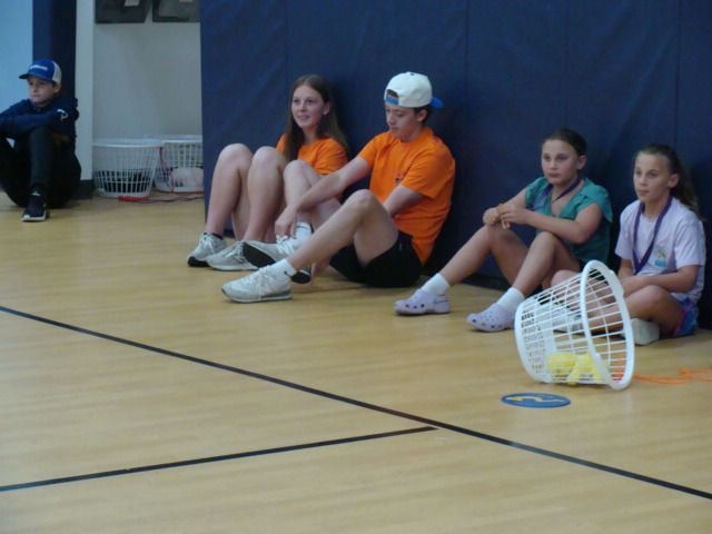 Students sitting on a gym floor beside a tipped-over white basket near a blue wall
