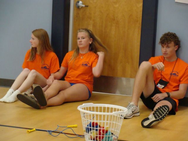 Three people in orange shirts sit on the floor with sports gear beside a basket in a gym.
