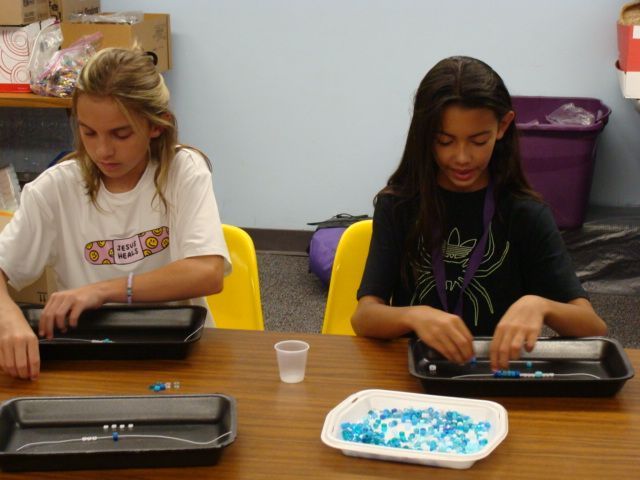 Two students using laptops at a table with a tray of blue beads in a classroom