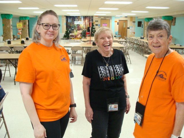 Three smiling volunteers in orange and black shirts standing in a cafeteria