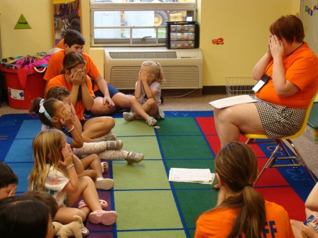 Children sitting on a classroom rug, listening to a seated teacher in an orange shirt.