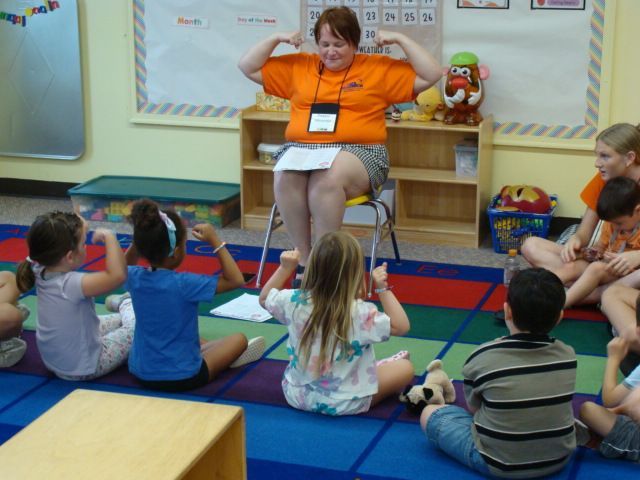 Teacher in orange shirt leading seated children in a classroom circle activity