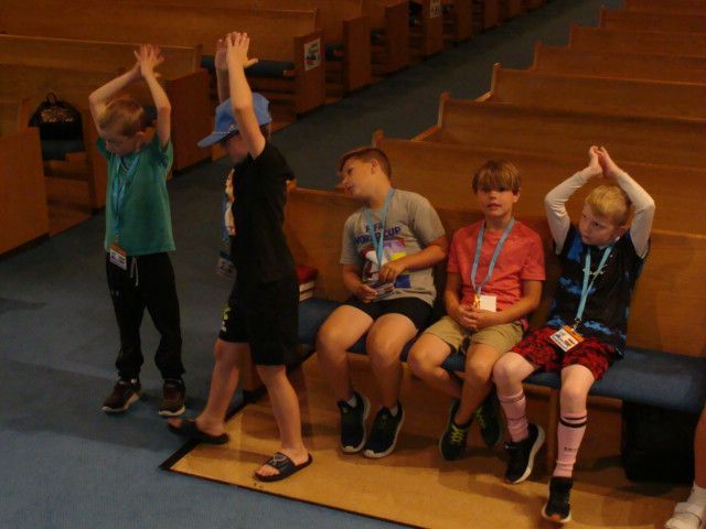 Children in a church pew doing stretches while seated, with one standing nearby raising arms
