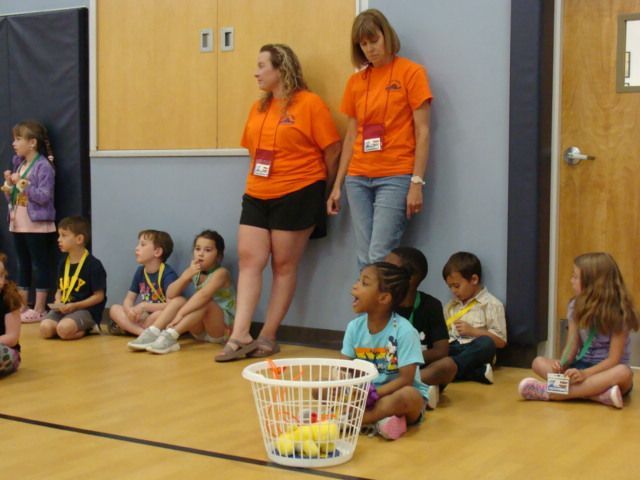 Children sitting on a gym floor with two adults in orange shirts nearby and a basket of balls in front