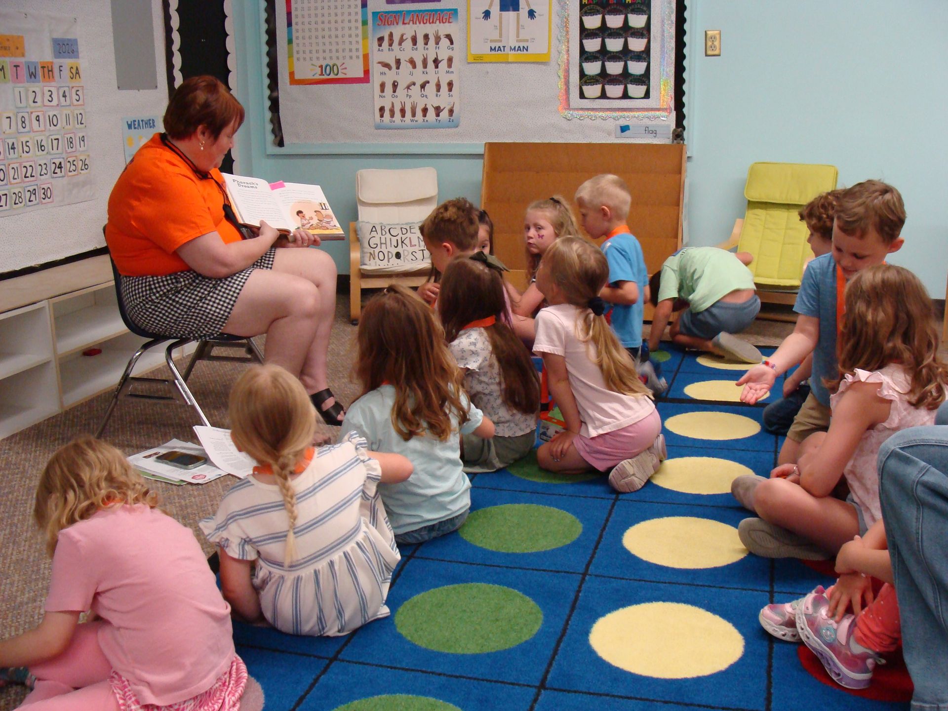 Teacher reading to a seated group of children on a blue and yellow classroom rug.