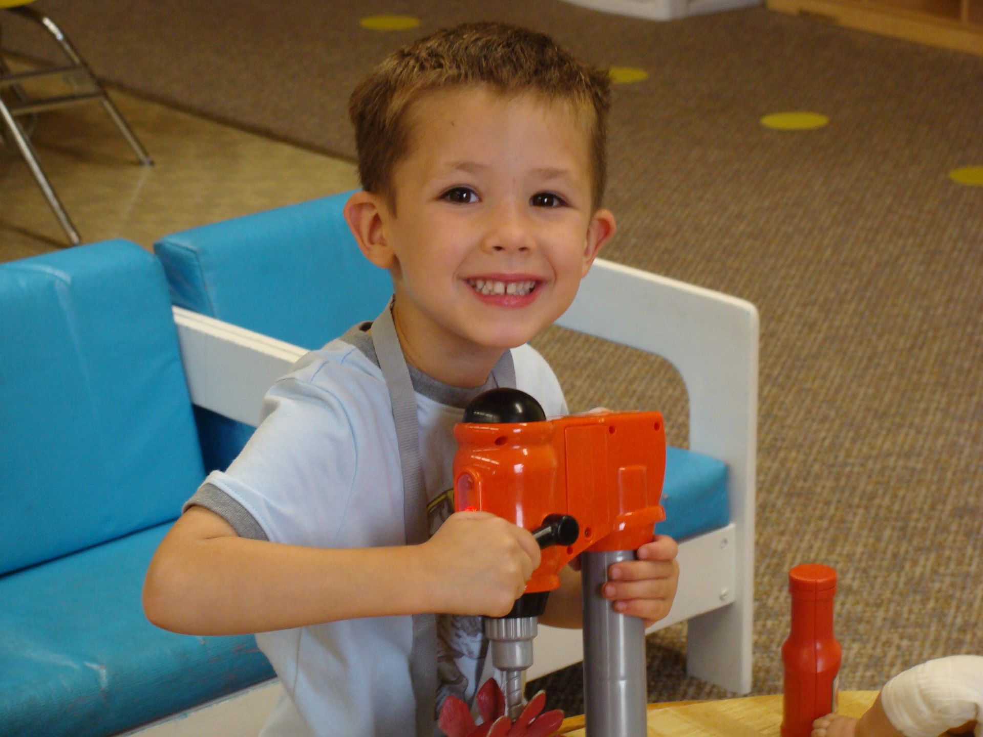 Smiling child holding an orange toy drill while sitting on a blue chair indoors