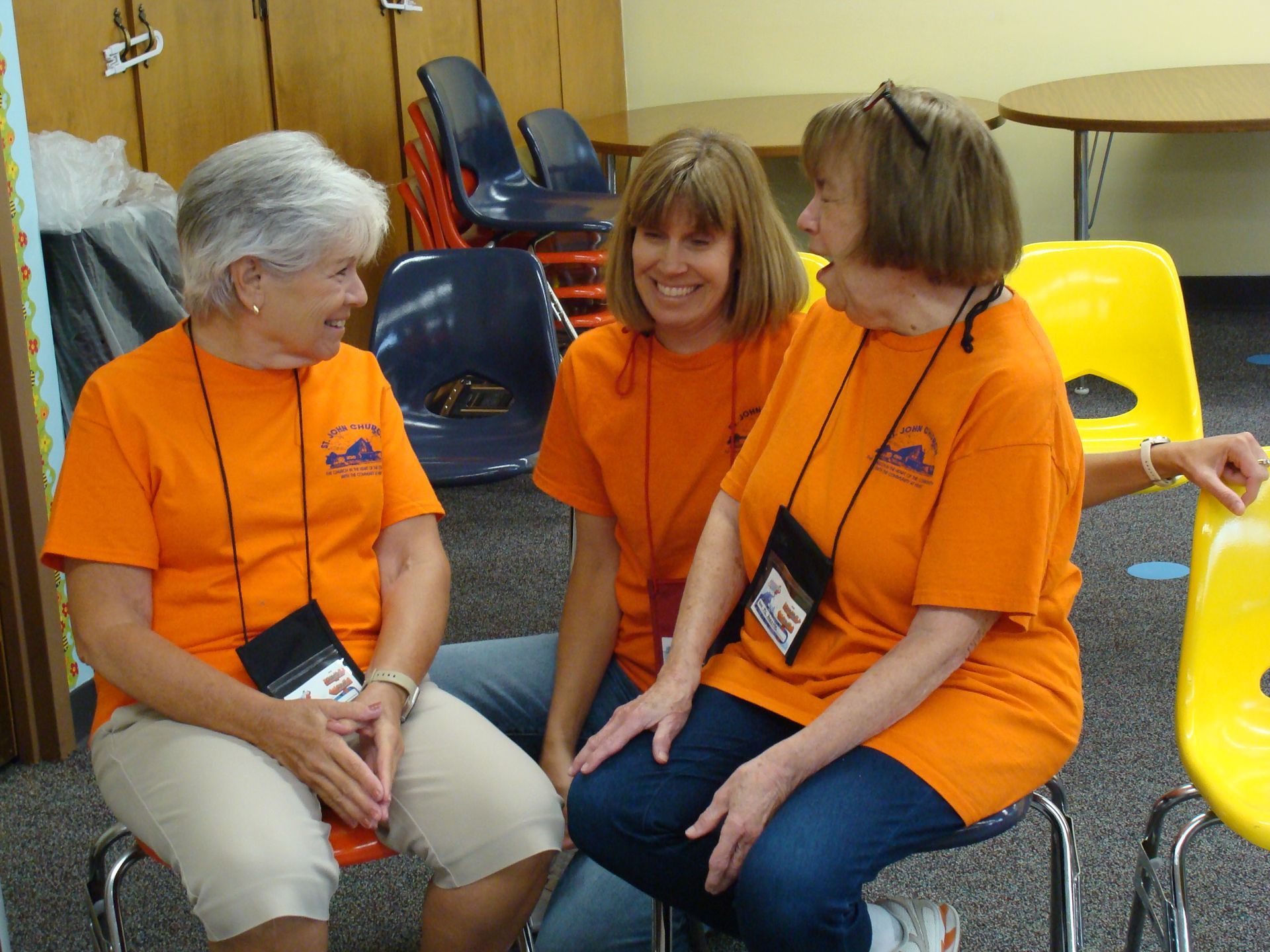 Three people in orange shirts sit chatting on chairs in a room with yellow and blue chairs.