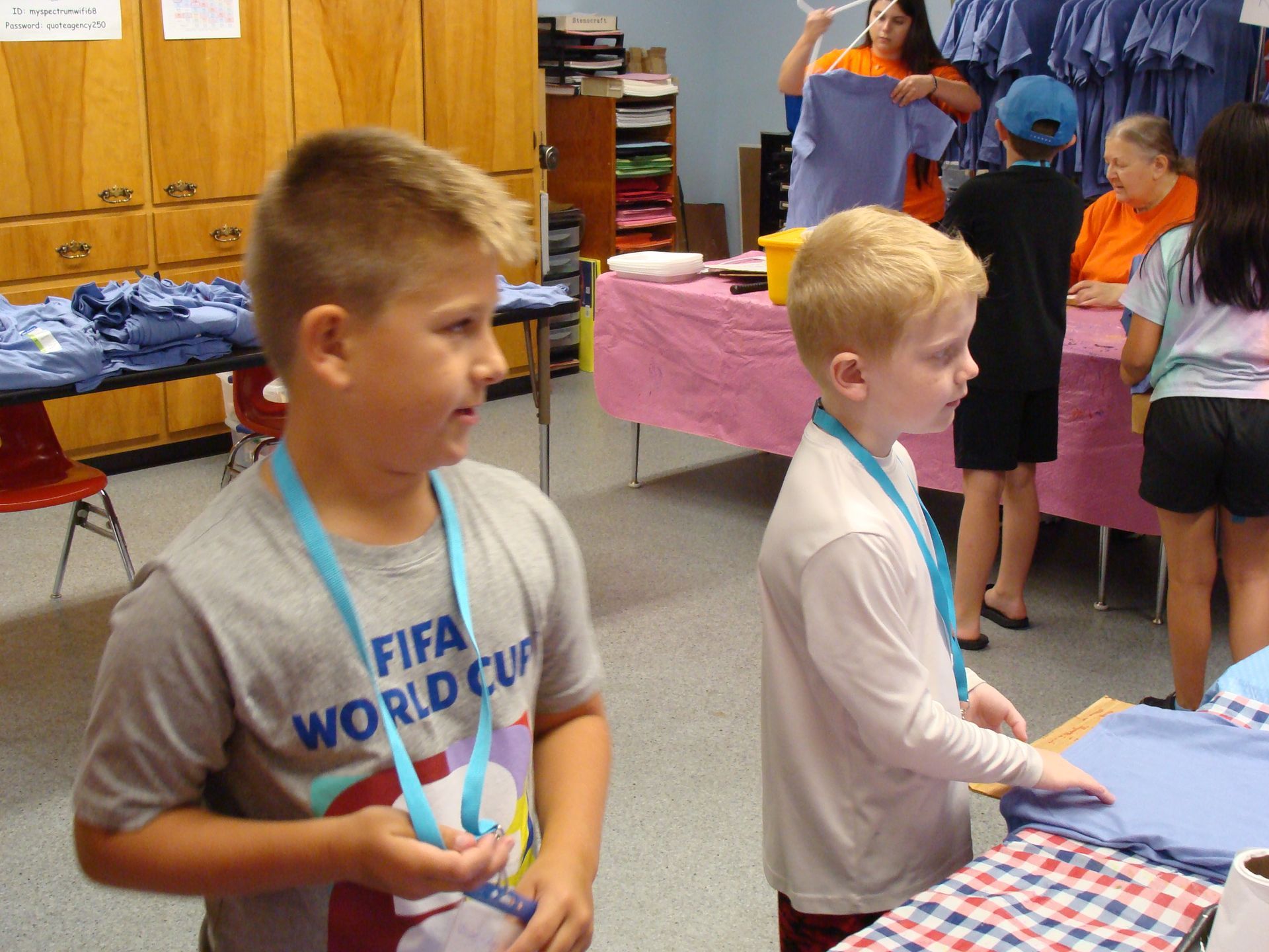 Children standing at a table in a classroom, looking at folded clothes and gift items