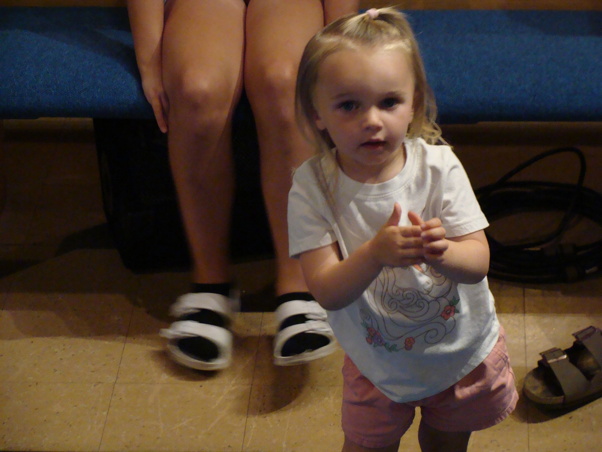 Toddler standing indoors beside seated adults, wearing a white shirt and pink shorts, with sandals on the floor.