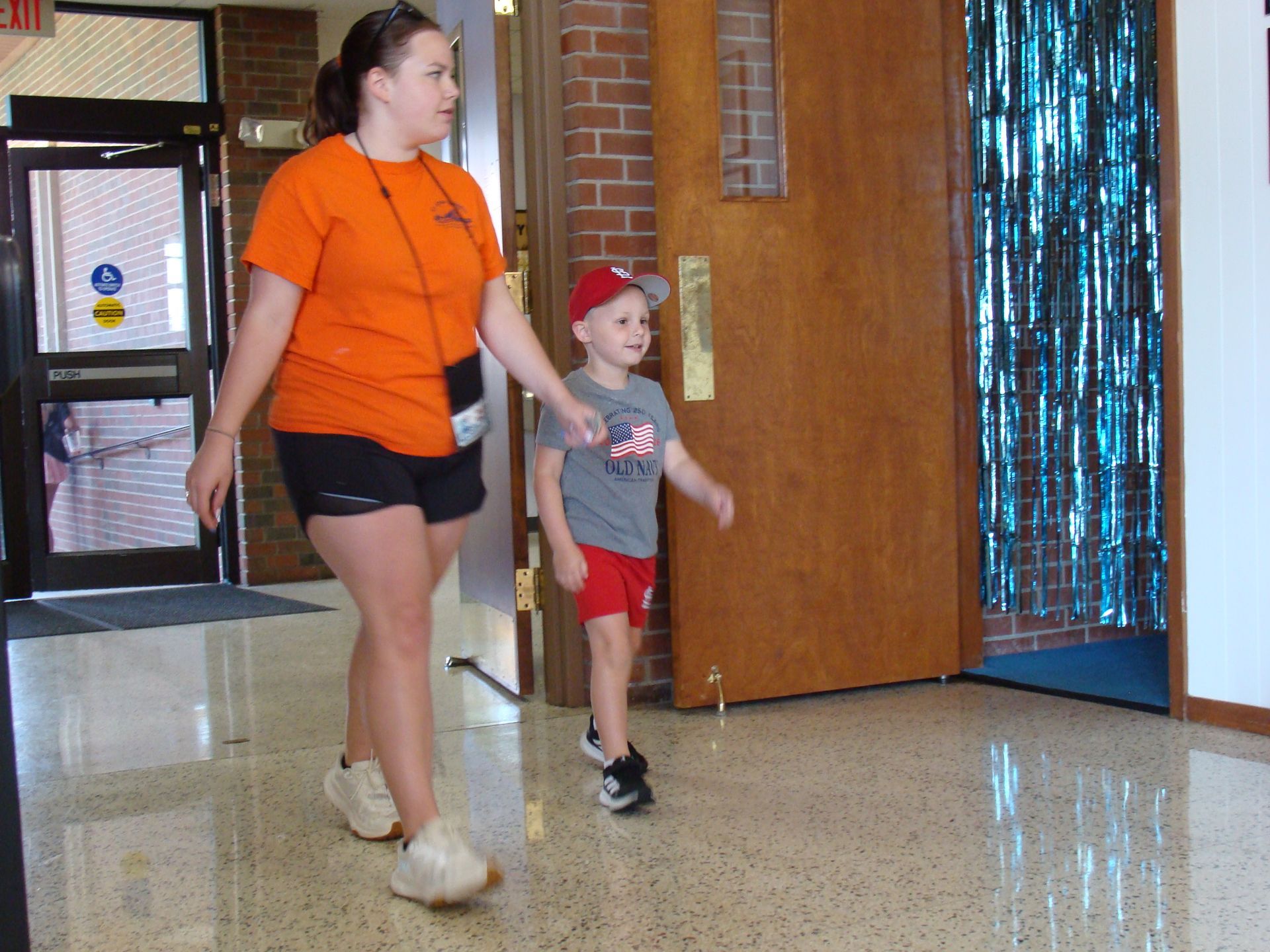 Adult and child walking indoors near a doorway, both wearing orange tops and shorts.