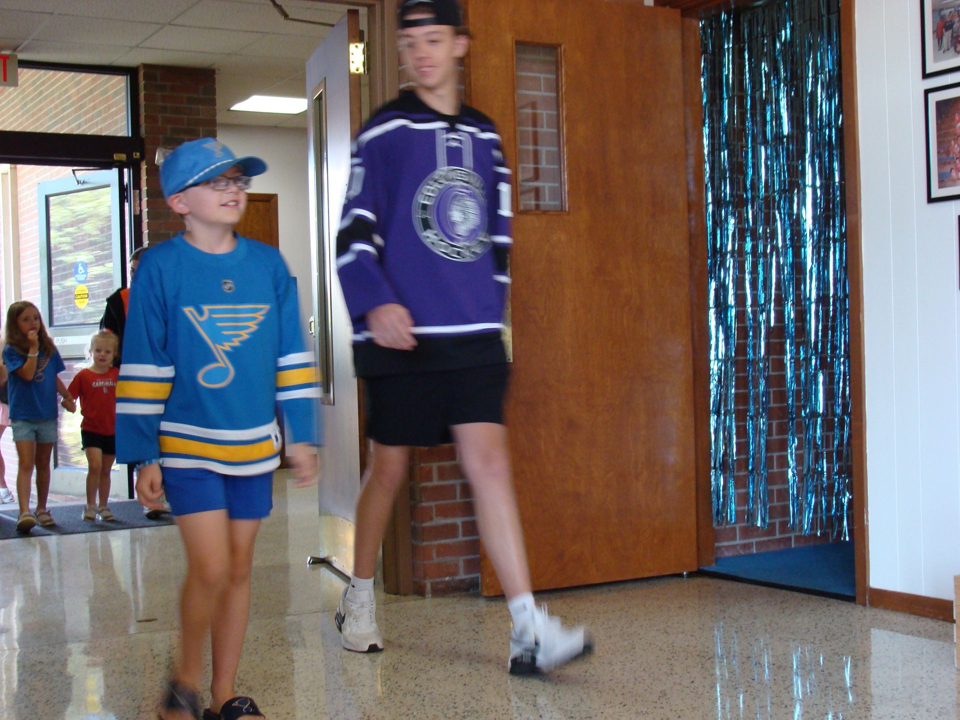 Two people in blue sports jerseys walk down a school hallway, one tall and one short.