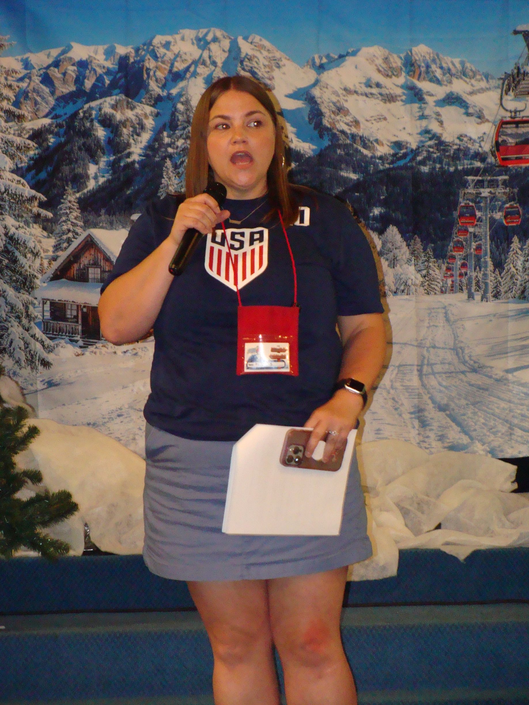 Woman in USA shirt holding a microphone and paper, standing before a snowy mountain backdrop.