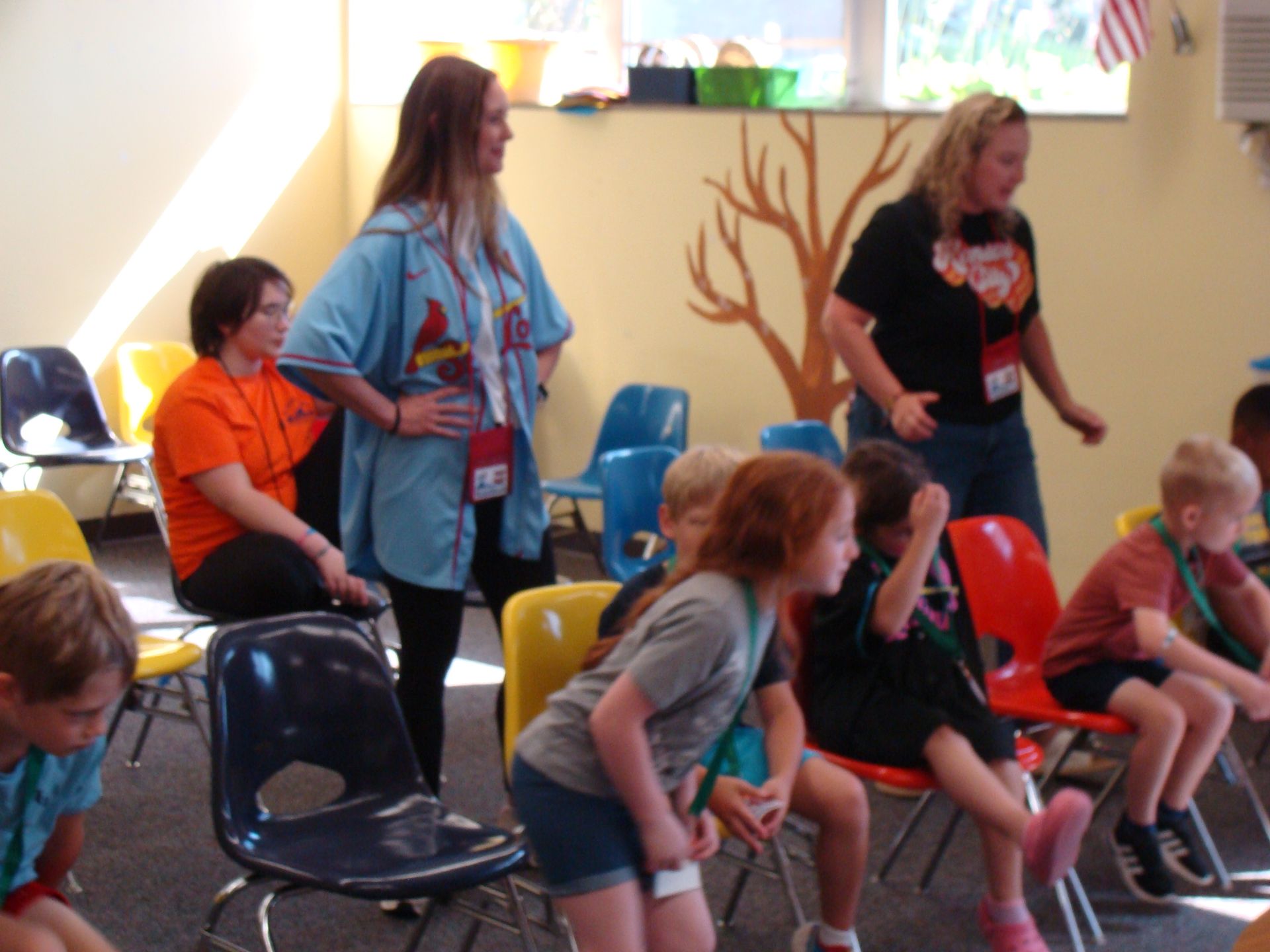 Children and adults doing a group exercise in a bright classroom, bending forward near chairs