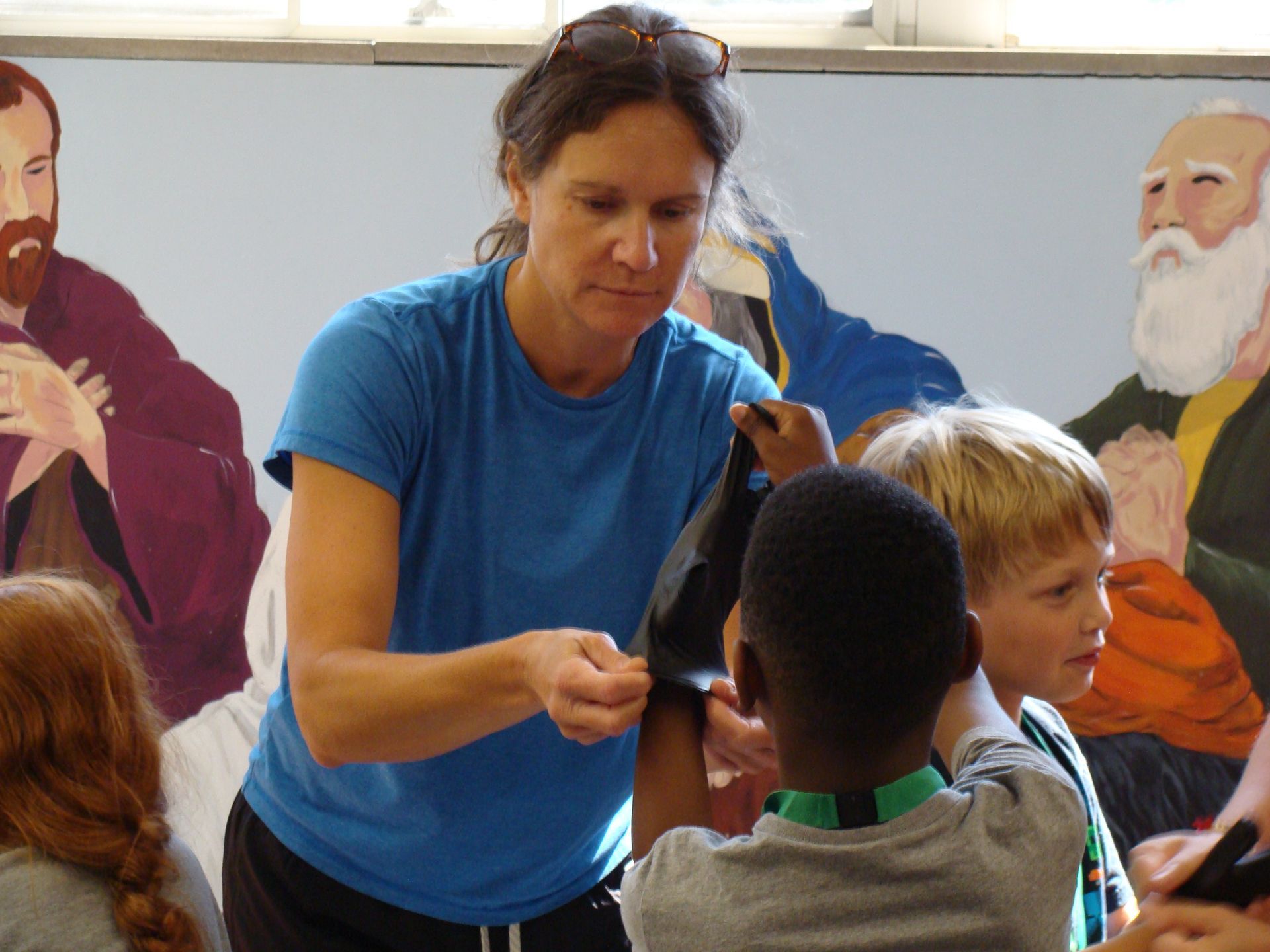 Teacher handing materials to children in a classroom with murals on the wall