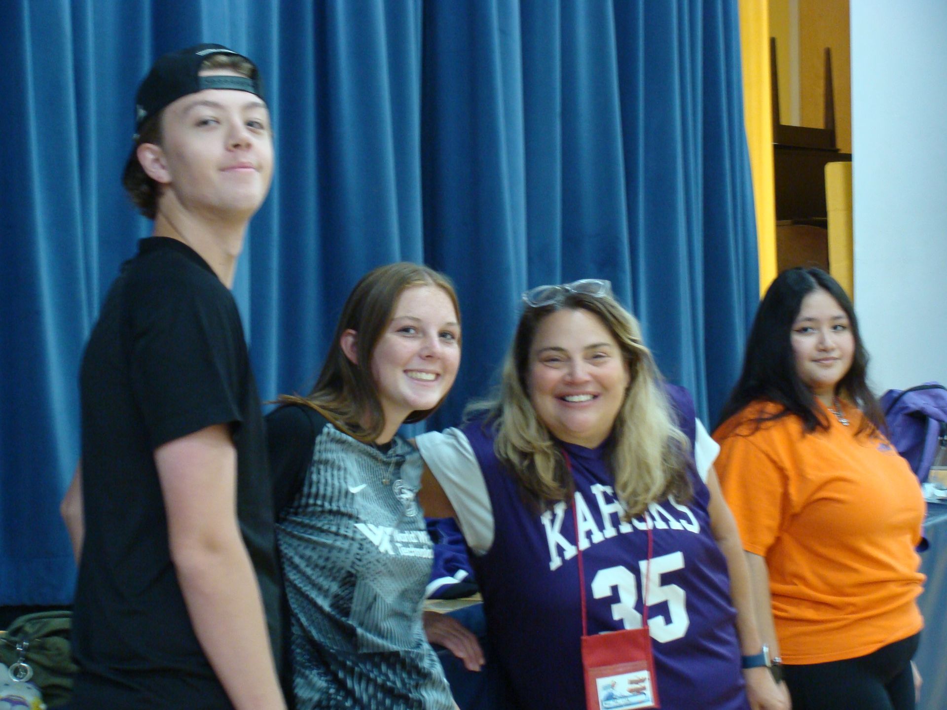 Four people posing in front of blue curtains, one wearing a purple Kappa jersey and one in an orange shirt