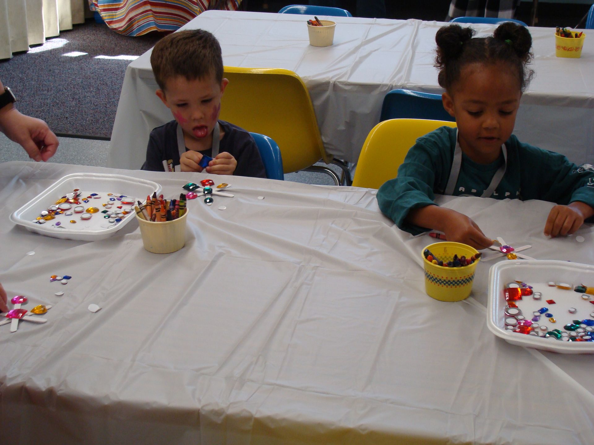 Children doing a craft activity at a table with beads, cups, and trays in a bright room