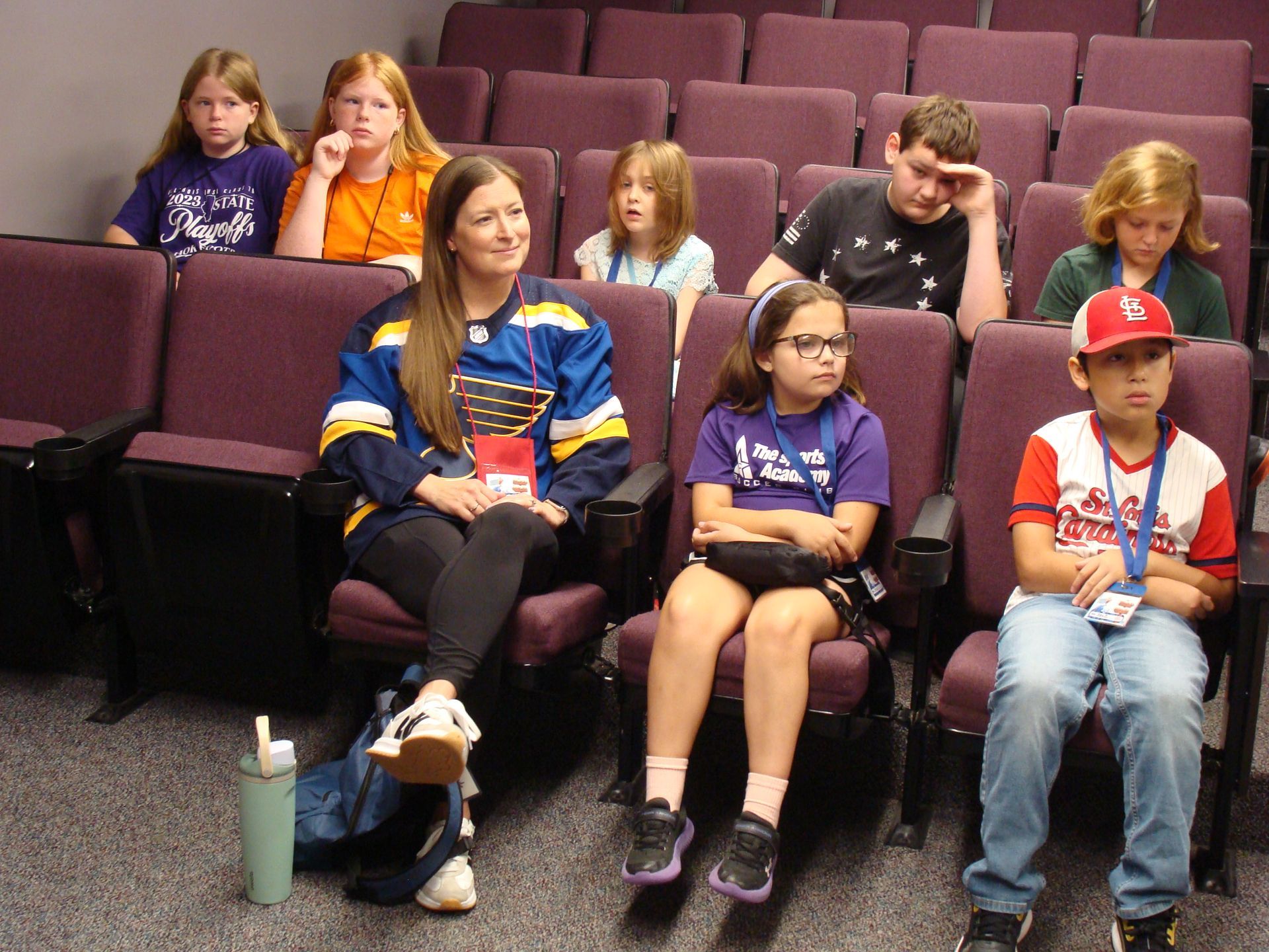 Children seated in a small auditorium, with one woman in front and a drink cup by her seat