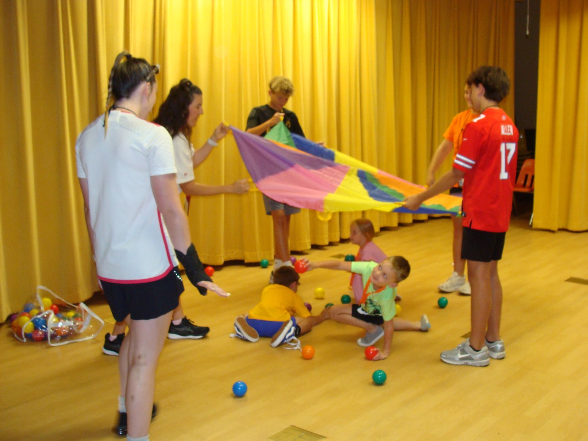 People holding a colorful parachute over children playing on a gym floor.