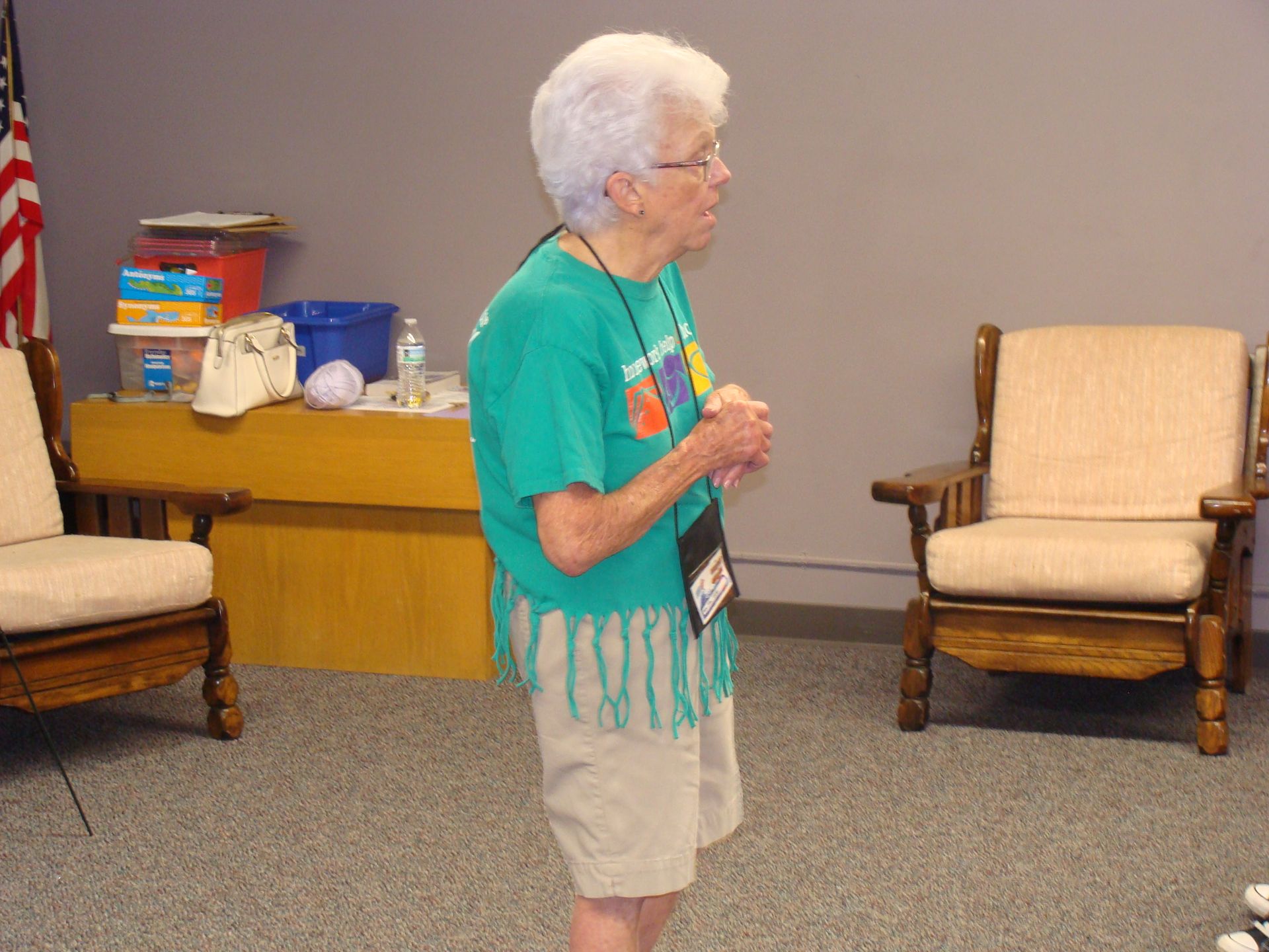 Person standing in a room, wearing a teal shirt and khaki shorts, near chairs and a side table with supplies