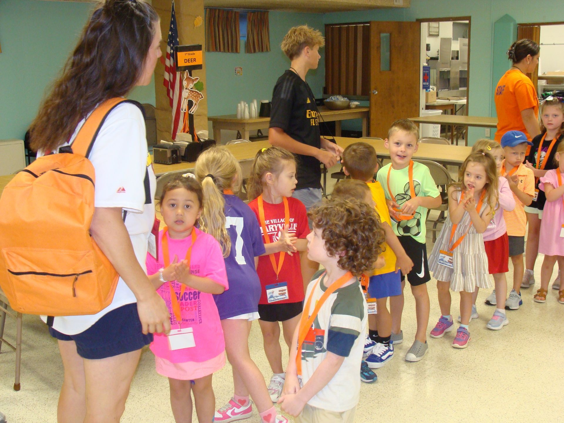Children lined up indoors, each holding hands while a woman with an orange backpack leads them.