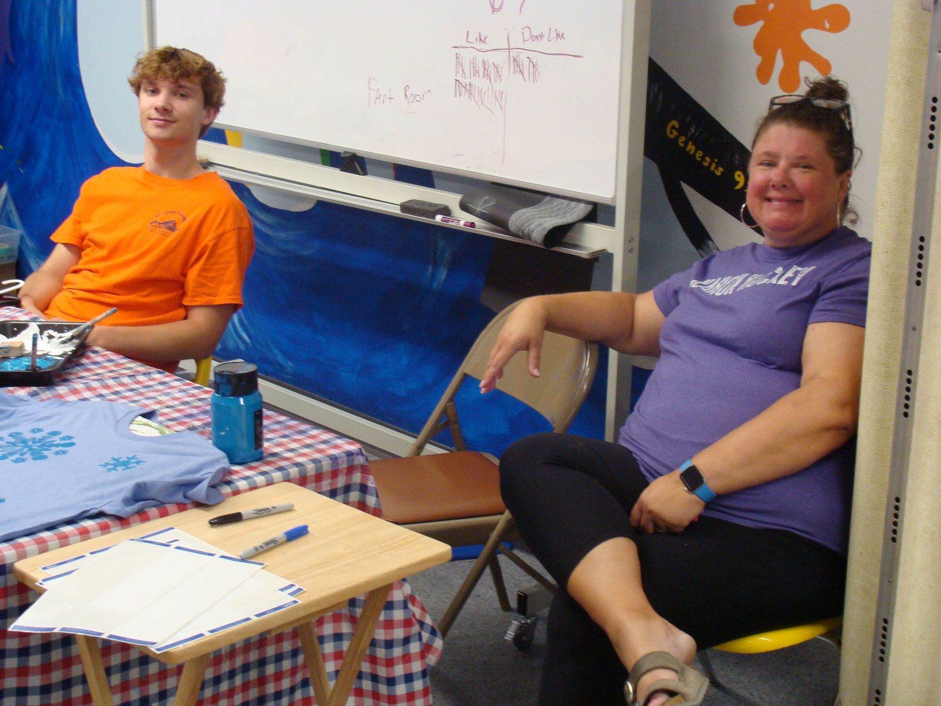 Two people seated at a table in a classroom, smiling with craft supplies and papers in front of them.
