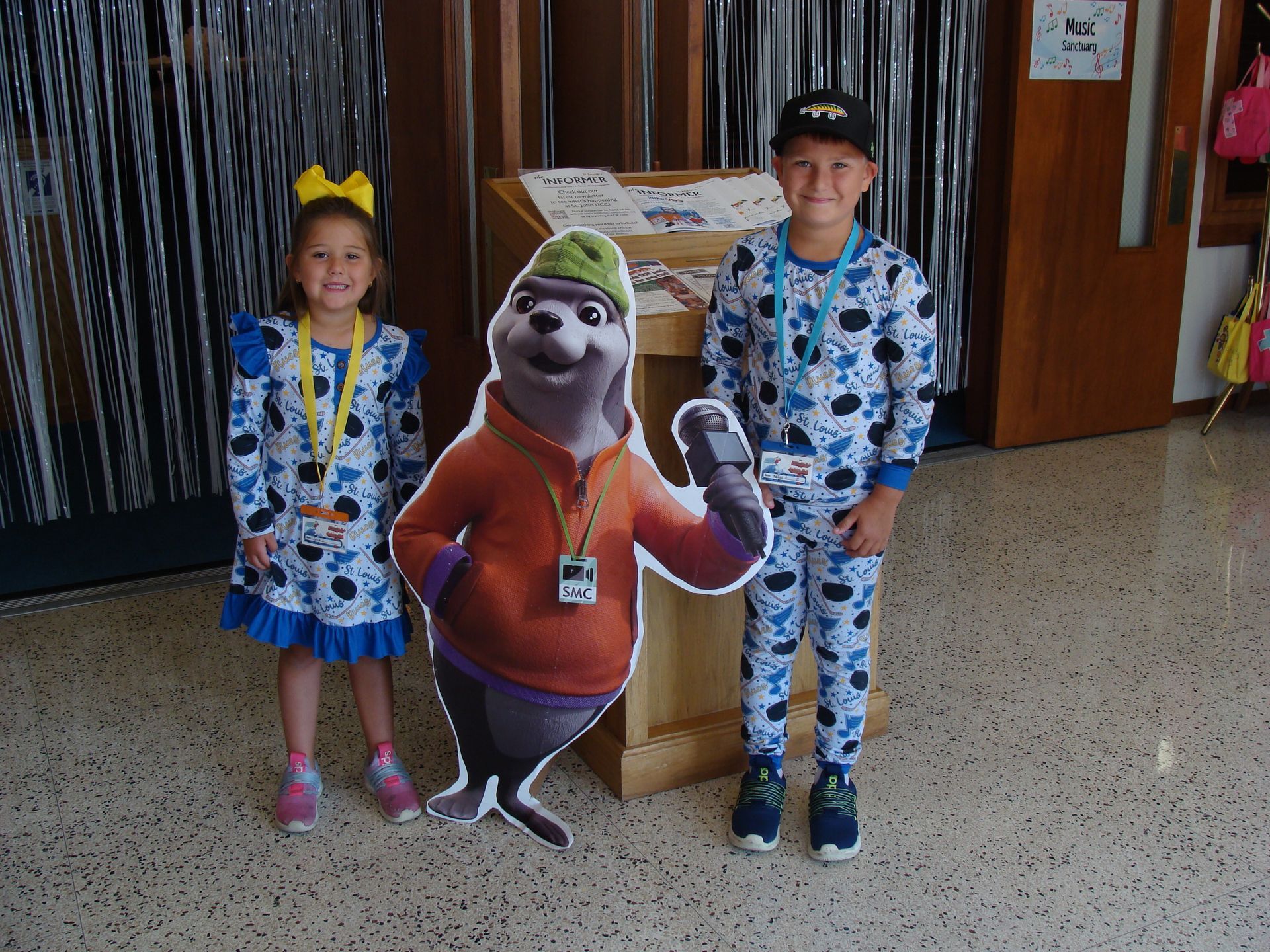 Two children stand beside a Shrek cutout indoors, one in a yellow bow and blue floral outfit.