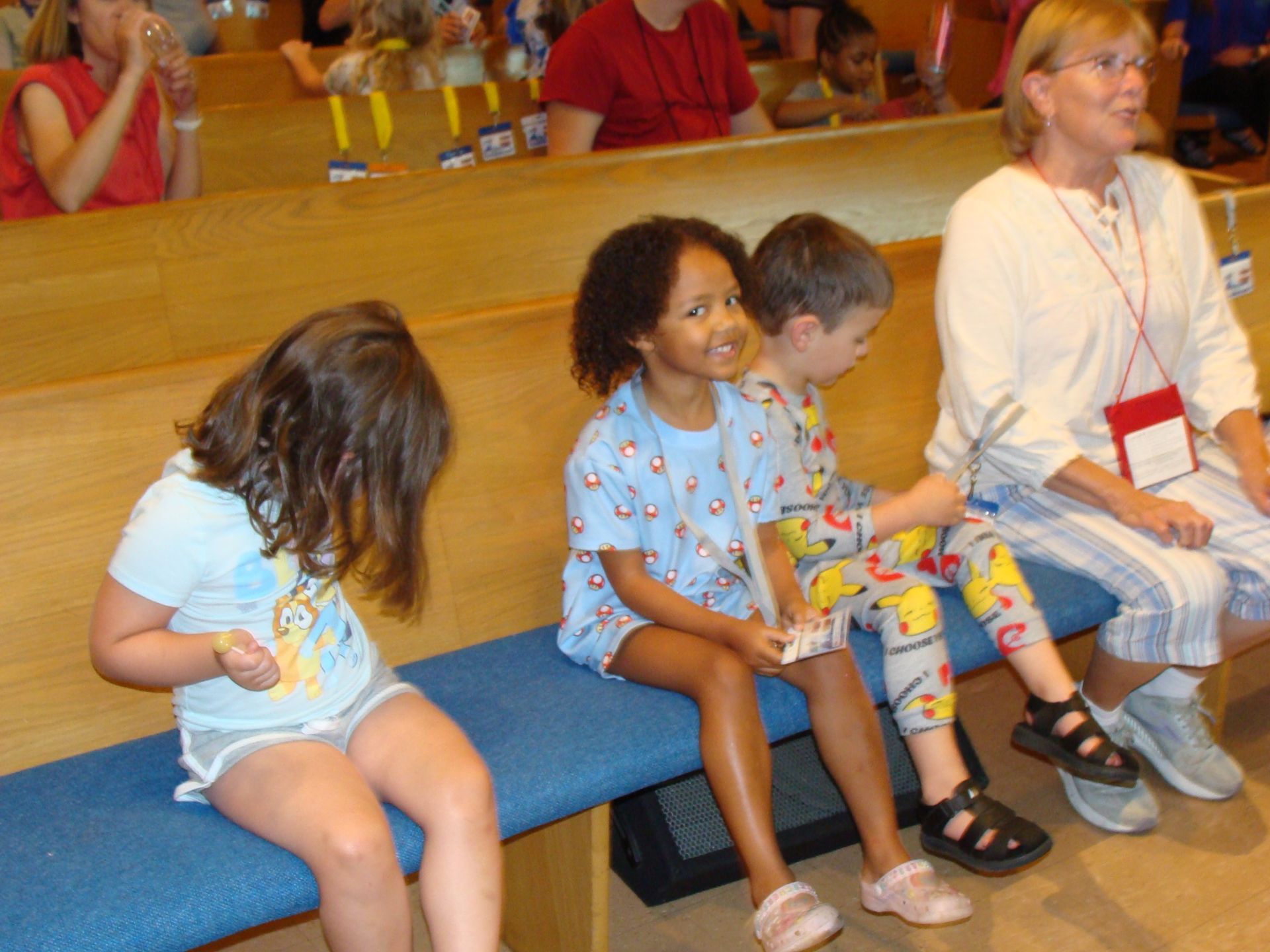 Children sitting on benches in a hall, looking down at papers and toys.