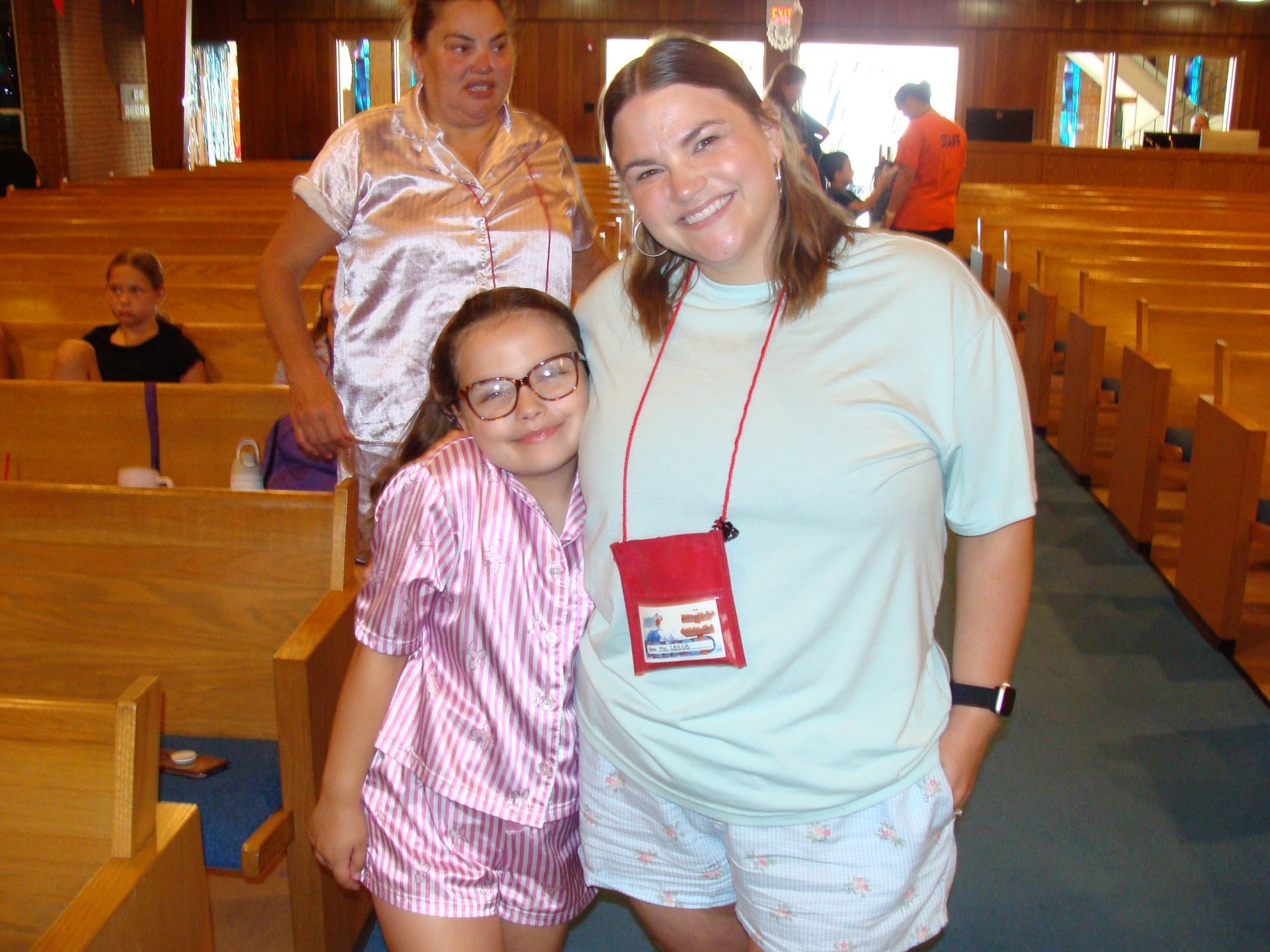 Two smiling people posing in a wooden church pew with a red lanyard badge and pews behind them