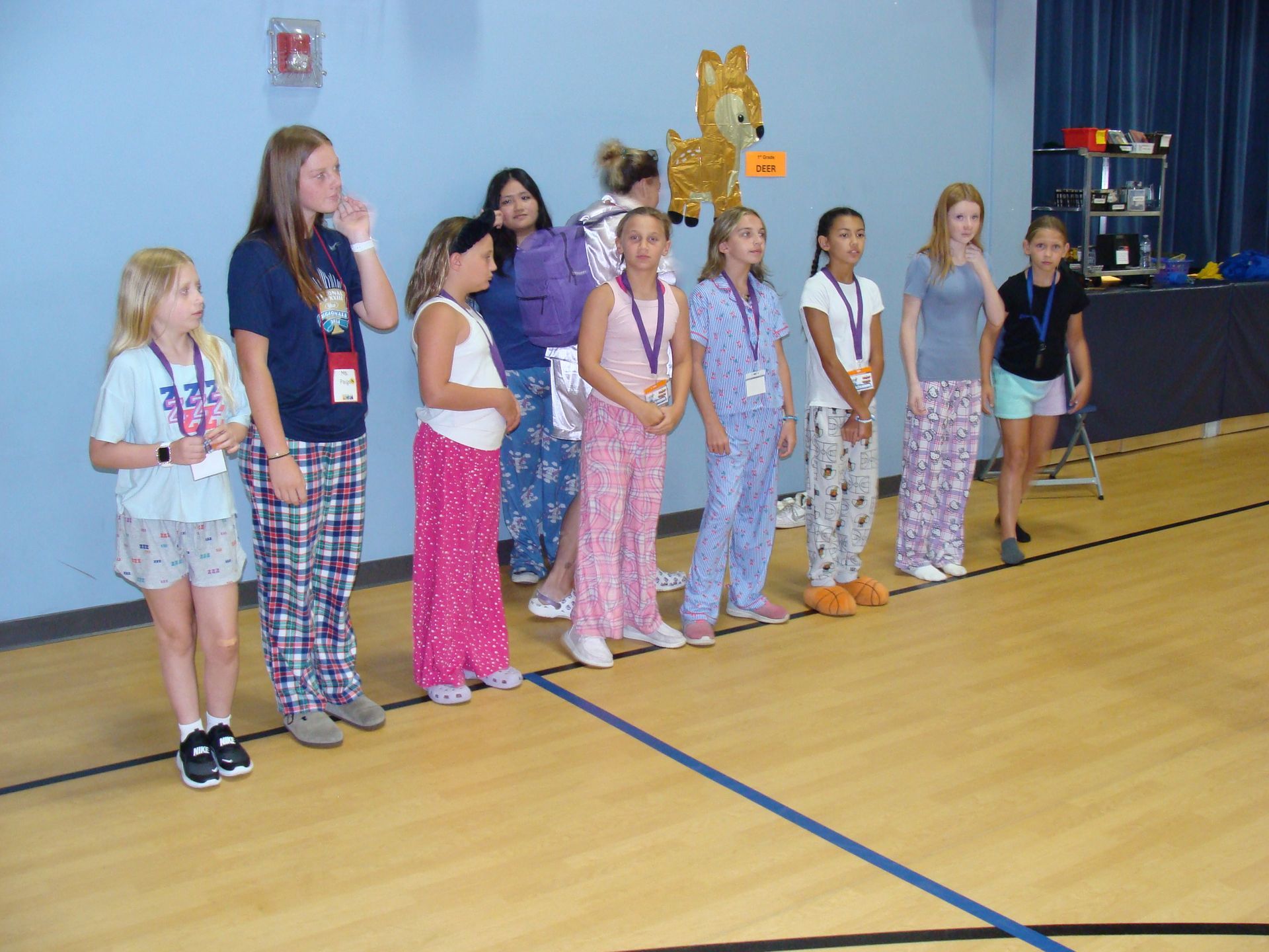 Children lined up indoors at an event, wearing lanyards and holding medals on a gym floor.