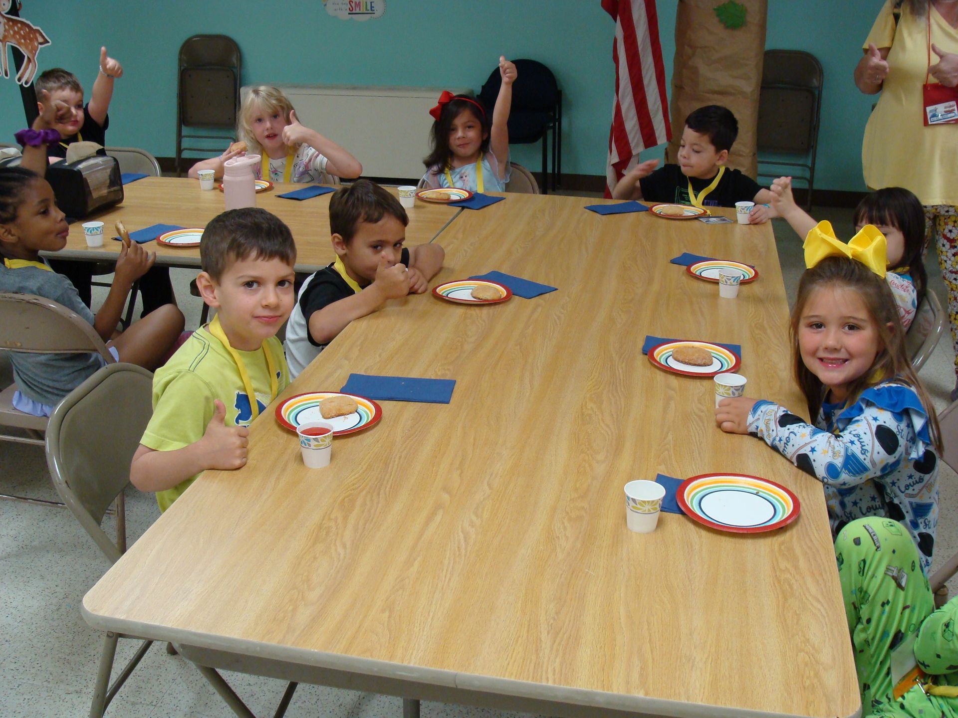 Children seated around a long table at a party, holding plates and cups in a classroom-like room.