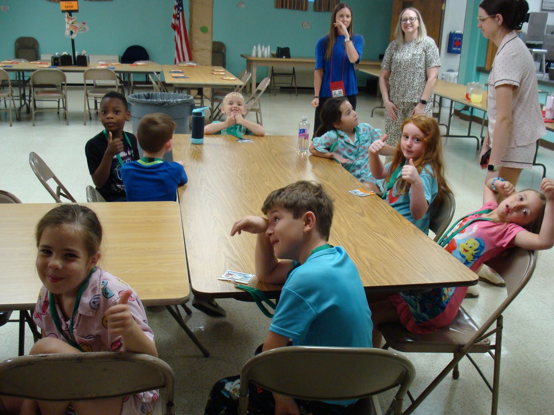 Children seated at folding tables in a classroom, with adults standing in the background.